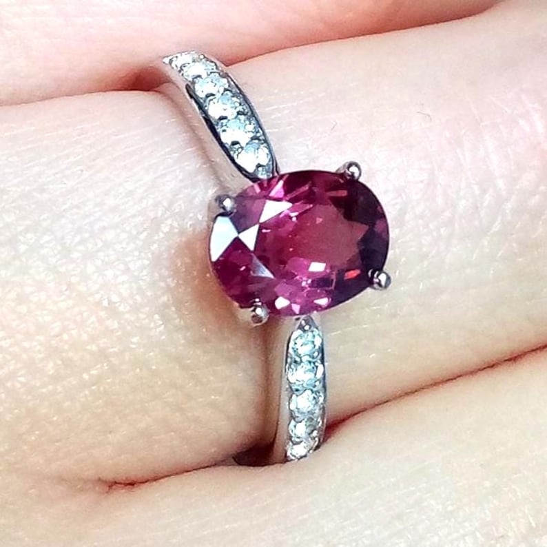 May include: A silver-toned ring featuring a large, oval-cut, deep pinkish-red gemstone held by four prongs. The band is accented with a row of small, clear, round-cut stones embedded along both sides of the central setting, creating a sparkling effect.