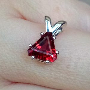 May include: A triangular-cut, deep red gemstone pendant set in a polished silver-toned metal prong setting. The pendant features a faceted surface that reflects light, held by a silver-toned bail at the top, showcasing a vibrant crimson hue.