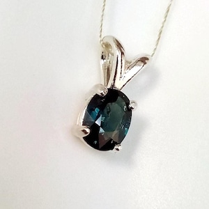 May include: A silver pendant necklace featuring an oval, dark teal gemstone. The gemstone is set in a silver bezel with a decorative bail. The necklace is displayed on a white background.