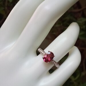 May include: A silver ring featuring a central oval-cut deep pink gemstone held by four prongs. The band is crafted from silver metal and is accented with small, clear, round-cut stones embedded along the shoulders on both sides of the center stone.