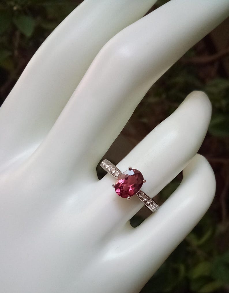 May include: A silver ring featuring a central oval-cut deep pink gemstone held by four prongs. The band is crafted from silver metal and is accented with small, clear, round-cut stones embedded along the shoulders on both sides of the center stone.