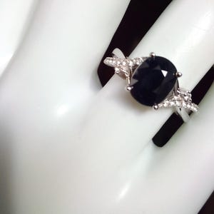Puede incluir: Un anillo plateado con una gran piedra preciosa ovalada de color azul oscuro como pieza central. La banda est&aacute; adornada con piedras m&aacute;s peque&ntilde;as y transparentes. El anillo se muestra sobre un fondo blanco, resaltando su dise&ntilde;o y detalles.
