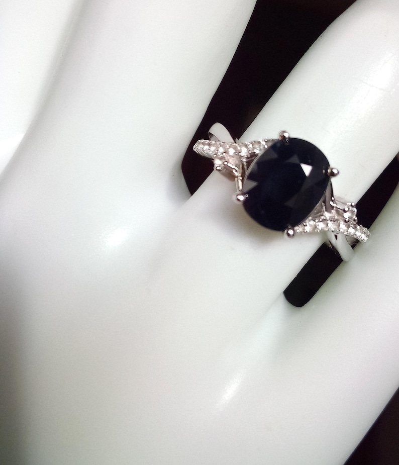 Puede incluir: Un anillo plateado con una gran piedra preciosa ovalada de color azul oscuro como pieza central. La banda est&aacute; adornada con piedras m&aacute;s peque&ntilde;as y transparentes. El anillo se muestra sobre un fondo blanco, resaltando su dise&ntilde;o y detalles.