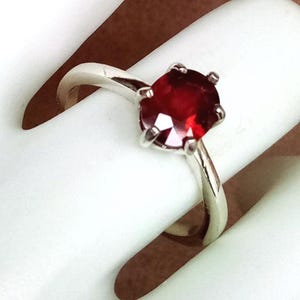May include: A silver ring featuring a round, faceted, deep red gemstone. The gemstone is held in place by four prongs. The ring band is smooth and polished.