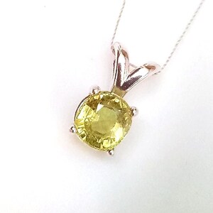 May include: A silver pendant necklace featuring a round, light yellow gemstone. The gemstone is held in place by four prongs and is suspended from a delicate silver chain. The pendant has a simple, elegant design.