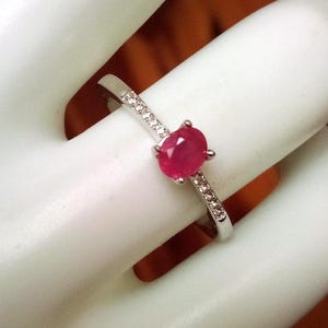 May include: A silver ring with a vibrant red oval gemstone at the center. The band is adorned with small, clear stones. The ring is displayed on a white surface, showcasing its elegant design and gemstone detail.