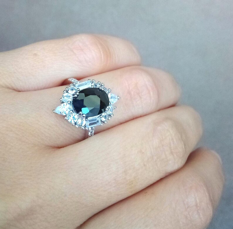 May include: A silver ring with a large, oval, dark teal gemstone at its centre, encircled by smaller, clear, baguette and pear-cut stones. The ring's delicate band enhances its glamorous appearance. A statement piece of jewellery.