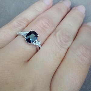 Puede incluir: Un anillo de plata con una piedra preciosa ovalada de color azul oscuro como pieza central. El anillo est&aacute; adornado con piedras preciosas m&aacute;s peque&ntilde;as y transparentes a ambos lados de la piedra principal. El anillo est&aacute; en una mano.