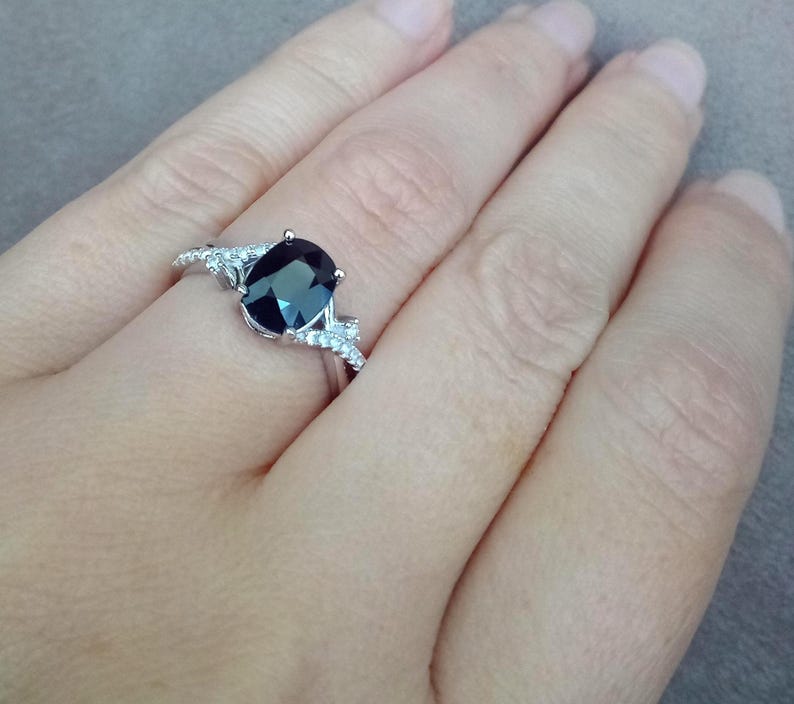 Puede incluir: Un anillo de plata con una piedra preciosa ovalada de color azul oscuro como pieza central. El anillo est&aacute; adornado con piedras preciosas m&aacute;s peque&ntilde;as y transparentes a ambos lados de la piedra principal. El anillo est&aacute; en una mano.