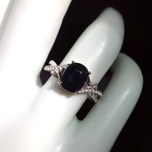 Puede incluir: Un anillo de plata con una piedra preciosa grande, ovalada y azul oscuro. La banda tiene un dise&ntilde;o dividido con peque&ntilde;as piedras de acento transparentes. El anillo se muestra sobre una superficie blanca.