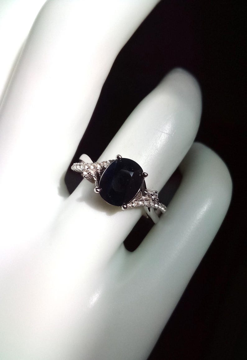 Puede incluir: Un anillo de plata con una piedra preciosa grande, ovalada y azul oscuro. La banda tiene un dise&ntilde;o dividido con peque&ntilde;as piedras de acento transparentes. El anillo se muestra sobre una superficie blanca.