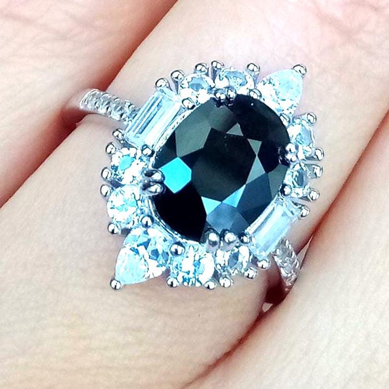 May include: A silver ring with a large, dark, faceted gemstone at its centre. The dark stone is encircled by smaller, clear, and baguette-cut stones, forming a halo. The ring's band is also embellished with smaller stones.