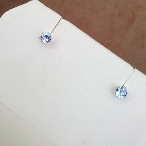 May include: A pair of small, light blue gemstone stud earrings. Each earring features a round, faceted gemstone set in a silver or white metal setting. The earrings are displayed on a white surface.