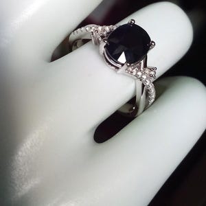 Puede incluir: Anillo plateado con una gran piedra negra de forma ovalada. La banda est&aacute; adornada con piedras preciosas claras m&aacute;s peque&ntilde;as. El anillo se muestra sobre una superficie blanca, resaltando su dise&ntilde;o elegante y detallado.