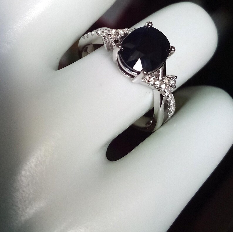 Puede incluir: Anillo plateado con una gran piedra negra de forma ovalada. La banda est&aacute; adornada con piedras preciosas claras m&aacute;s peque&ntilde;as. El anillo se muestra sobre una superficie blanca, resaltando su dise&ntilde;o elegante y detallado.
