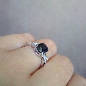 Puede incluir: Un anillo plateado con una gran piedra preciosa azul oscuro como pieza central. El anillo tiene un dise&ntilde;o delicado con piedras m&aacute;s peque&ntilde;as y claras que acent&uacute;an la banda. El anillo se muestra en el dedo de una persona, mostrando su estilo elegante y sofisticado.
