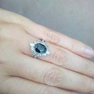 May include: A silver ring with a large, oval-cut dark green gemstone, encircled by smaller, clear gemstones. The ring has a halo design with baguette and pear-shaped accents, creating a sparkling, elegant appearance. The ring is displayed on a person's finger.