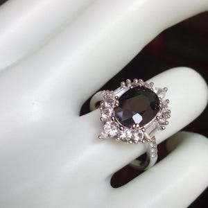 May include: A silver ring featuring a large, oval, dark grey gemstone at its centre, encircled by smaller, clear, rectangular and round stones. The ring is set on a white surface.