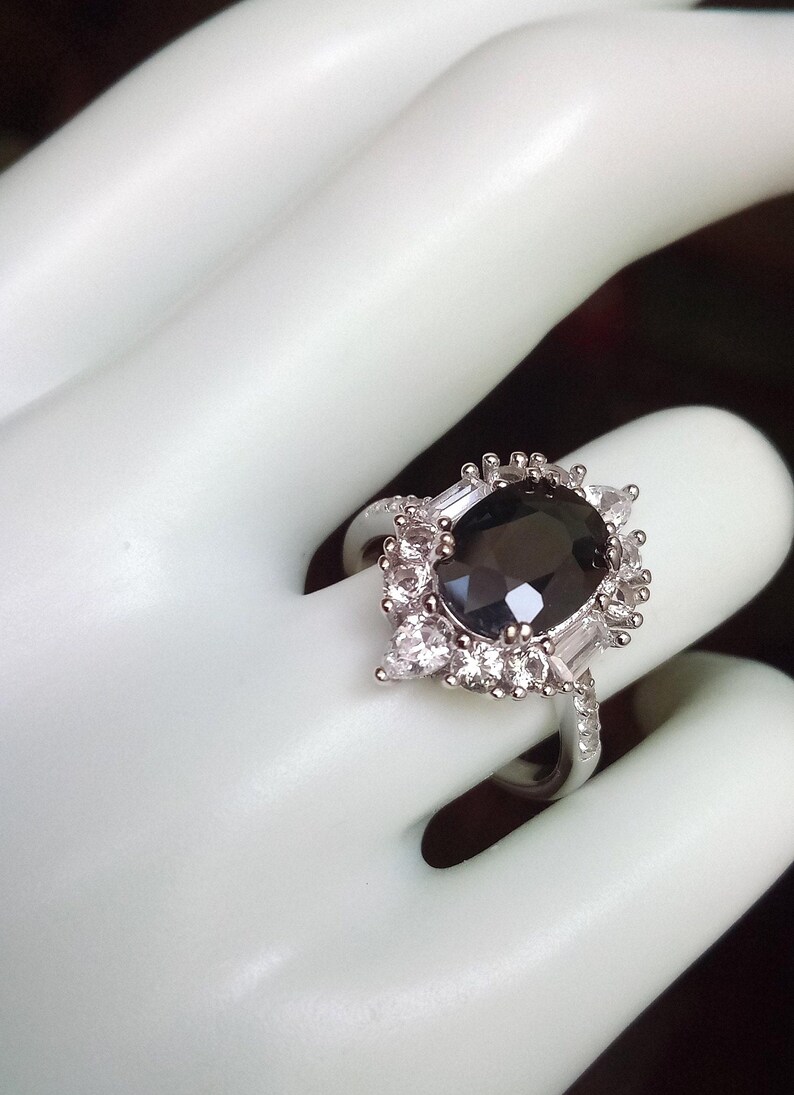 May include: A silver ring featuring a large, oval, dark grey gemstone at its centre, encircled by smaller, clear, rectangular and round stones. The ring is set on a white surface.