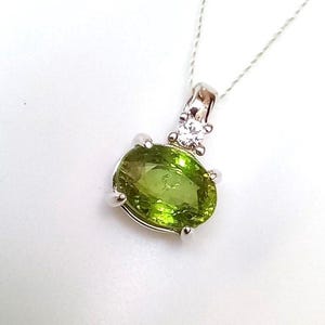 May include: A silver pendant necklace featuring a large, oval-cut, lime green peridot gemstone held by four prongs. Above the main stone, a smaller, round, clear white gemstone is set in a silver bail, suspended from a delicate silver chain.