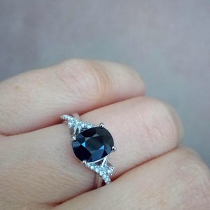 Puede incluir: Un anillo de plata con una gran gema ovalada de color azul oscuro. La banda del anillo tiene un dise&ntilde;o dividido con peque&ntilde;as piedras claras. La gema azul oscuro es el punto focal, engastada en un engaste de garras cl&aacute;sico.