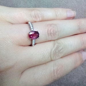 May include: A silver-toned ring featuring a central oval-cut deep pink gemstone held in a prong setting, with a slender band accented by small, clear, round-cut stones embedded along the sides of the shank, creating a refined and polished appearance.