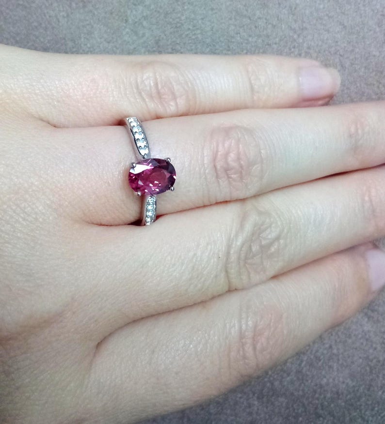 May include: A silver-toned ring featuring a central oval-cut deep pink gemstone held in a prong setting, with a slender band accented by small, clear, round-cut stones embedded along the sides of the shank, creating a refined and polished appearance.