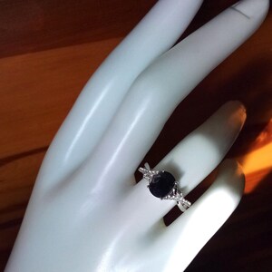 Puede incluir: Un anillo plateado con una gran piedra preciosa ovalada de color azul oscuro. El anillo tiene un dise&ntilde;o de banda dividida con piedras de acento m&aacute;s peque&ntilde;as y claras. El anillo se muestra en una mano blanca. El dise&ntilde;o es elegante.