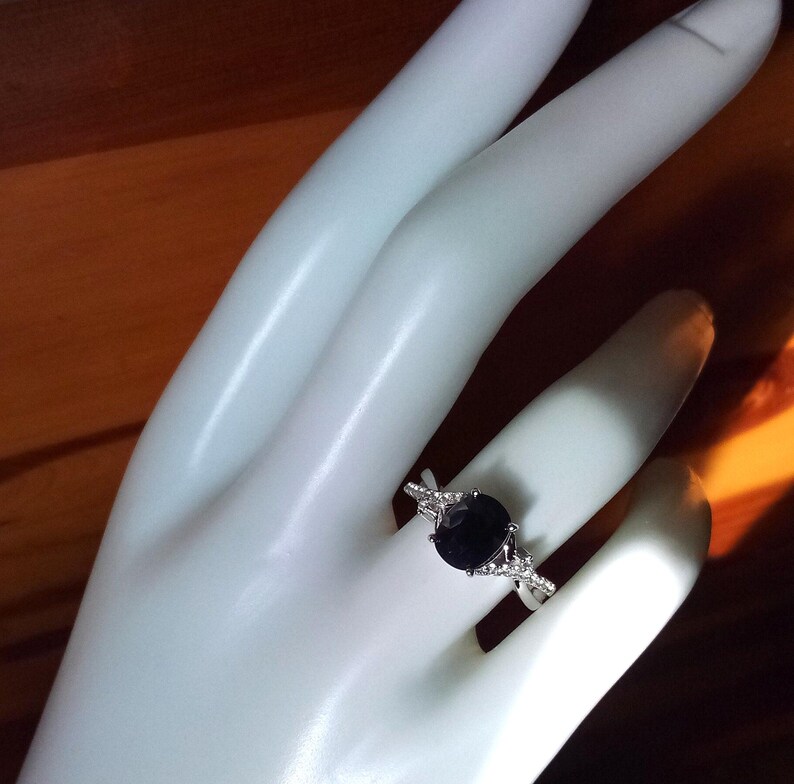 Puede incluir: Un anillo plateado con una gran piedra preciosa ovalada de color azul oscuro. El anillo tiene un dise&ntilde;o de banda dividida con piedras de acento m&aacute;s peque&ntilde;as y claras. El anillo se muestra en una mano blanca. El dise&ntilde;o es elegante.