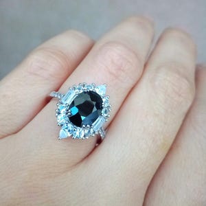 May include: A silver ring with a large, oval-cut, dark gemstone encircled by smaller, light-coloured stones. The ring's band is also decorated with small, sparkling accents. The ring is set against a neutral background.