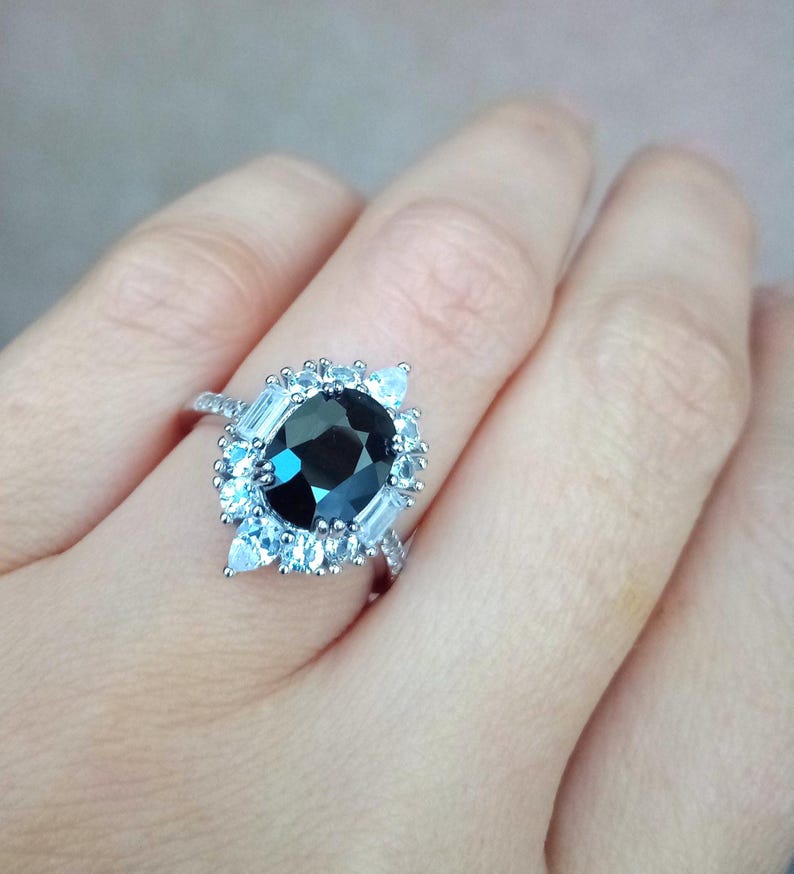 May include: A silver ring with a large, oval-cut, dark gemstone encircled by smaller, light-coloured stones. The ring's band is also decorated with small, sparkling accents. The ring is set against a neutral background.