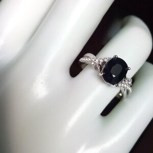 Puede incluir: Anillo de plata con una gran piedra negra de corte ovalado. La banda presenta un dise&ntilde;o retorcido con piedras preciosas m&aacute;s peque&ntilde;as y transparentes. El anillo se muestra sobre un fondo blanco.