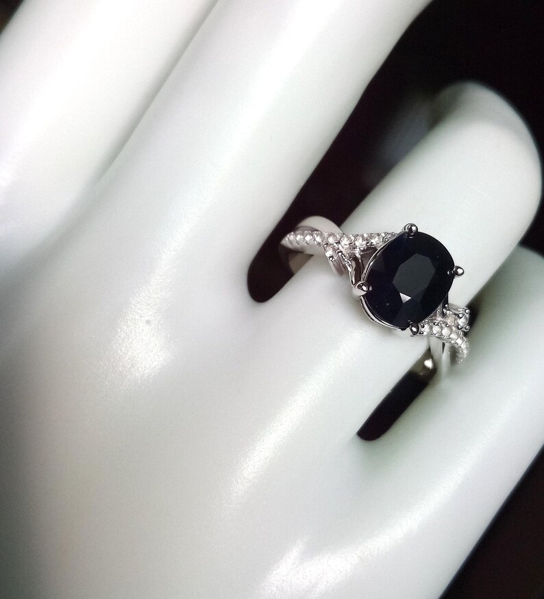 Puede incluir: Anillo de plata con una gran piedra negra de corte ovalado. La banda presenta un dise&ntilde;o retorcido con piedras preciosas m&aacute;s peque&ntilde;as y transparentes. El anillo se muestra sobre un fondo blanco.