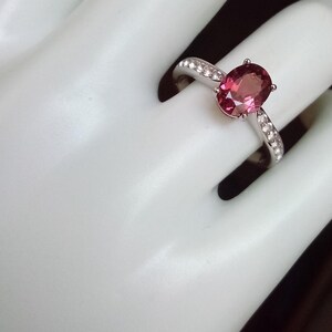 May include: A silver ring featuring a central oval-cut pink gemstone held by four prongs. The band is crafted from a polished silver-toned metal and is accented with a row of small, clear, round-cut stones embedded along each side of the shank.