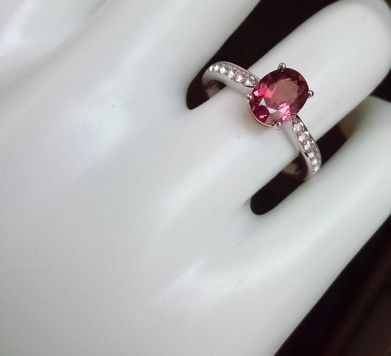 May include: A silver ring featuring a central oval-cut pink gemstone held by four prongs. The band is crafted from a polished silver-toned metal and is accented with a row of small, clear, round-cut stones embedded along each side of the shank.