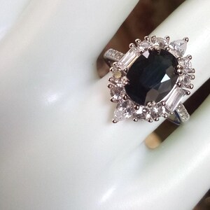 May include: A silver ring with a large, dark blue gemstone at the centre, encircled by smaller, clear stones. The ring has a halo design with a cluster of clear stones around the central stone. The band is also decorated with smaller clear stones.