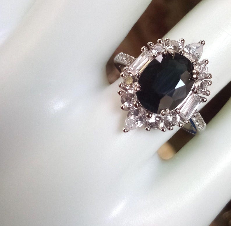 May include: A silver ring with a large, dark blue gemstone at the centre, encircled by smaller, clear stones. The ring has a halo design with a cluster of clear stones around the central stone. The band is also decorated with smaller clear stones.