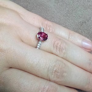 May include: A silver ring featuring a central oval-cut deep pink gemstone held in a four-prong setting. The band is crafted from polished silver and is accented with a row of small, clear, round-cut stones embedded along the shank on both sides.