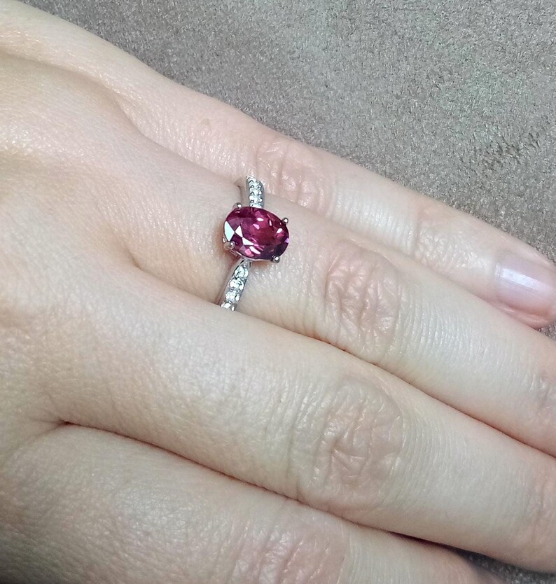 May include: A silver ring featuring a central oval-cut deep pink gemstone held in a four-prong setting. The band is crafted from polished silver and is accented with a row of small, clear, round-cut stones embedded along the shank on both sides.