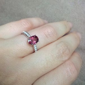 May include: A silver ring featuring a central oval-cut deep pink gemstone held by four prongs. The band is crafted from polished silver and is accented with small, clear, round-cut stones embedded along the shoulders on both sides of the center stone.