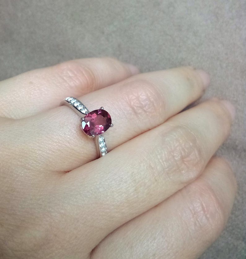 May include: A silver ring featuring a central oval-cut deep pink gemstone held by four prongs. The band is crafted from polished silver and is accented with small, clear, round-cut stones embedded along the shoulders on both sides of the center stone.