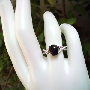 Puede incluir: Un anillo plateado con una piedra preciosa oscura, grande y ovalada. El anillo tiene una banda dividida con peque&ntilde;as piedras claras de acento. El anillo se muestra en una mano blanca, con un fondo verde borroso.