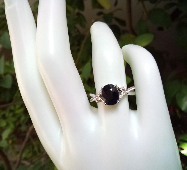 Puede incluir: Un anillo plateado con una piedra preciosa oscura, grande y ovalada. El anillo tiene una banda dividida con peque&ntilde;as piedras claras de acento. El anillo se muestra en una mano blanca, con un fondo verde borroso.