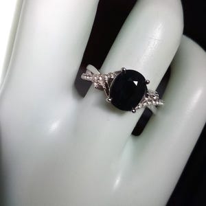 Puede incluir: Un anillo plateado con una gran piedra negra de corte ovalado. La banda est&aacute; adornada con piedras preciosas m&aacute;s peque&ntilde;as y claras a ambos lados de la piedra principal, creando un efecto brillante. El anillo se muestra sobre una superficie blanca.