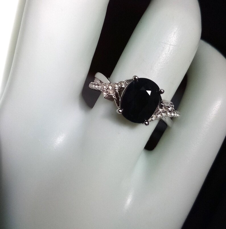 Puede incluir: Un anillo plateado con una gran piedra negra de corte ovalado. La banda est&aacute; adornada con piedras preciosas m&aacute;s peque&ntilde;as y claras a ambos lados de la piedra principal, creando un efecto brillante. El anillo se muestra sobre una superficie blanca.