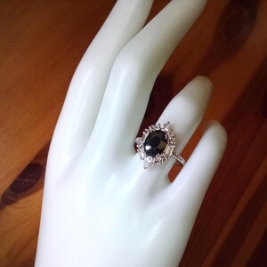 May include: A silver ring with a large, oval black gemstone, encircled by smaller, clear stones. The ring has an intricate setting and is shown on a white hand, set against a wooden backdrop.