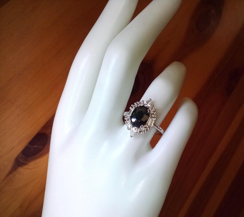 May include: A silver ring with a large, oval black gemstone, encircled by smaller, clear stones. The ring has an intricate setting and is shown on a white hand, set against a wooden backdrop.