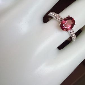 May include: A silver-toned ring featuring a central oval-cut pink gemstone held by four prongs. The band is accented with small, clear, round-cut stones embedded along the shoulders on both sides of the main setting, creating a polished look.