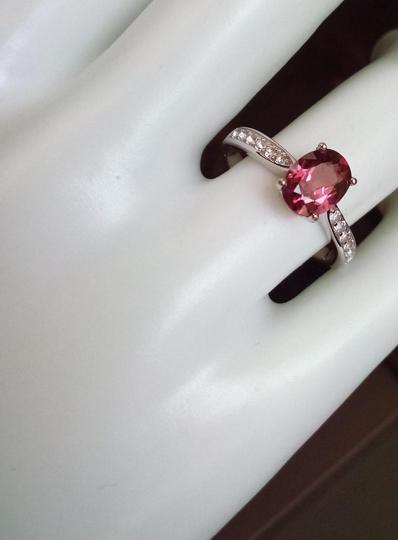 May include: A silver-toned ring featuring a central oval-cut pink gemstone held by four prongs. The band is accented with small, clear, round-cut stones embedded along the shoulders on both sides of the main setting, creating a polished look.