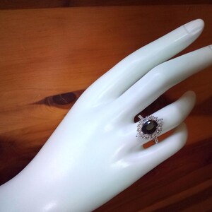 May include: A silver ring featuring an oval black gemstone, encircled by smaller, clear stones. The ring is displayed on a white hand, set against a wooden backdrop.